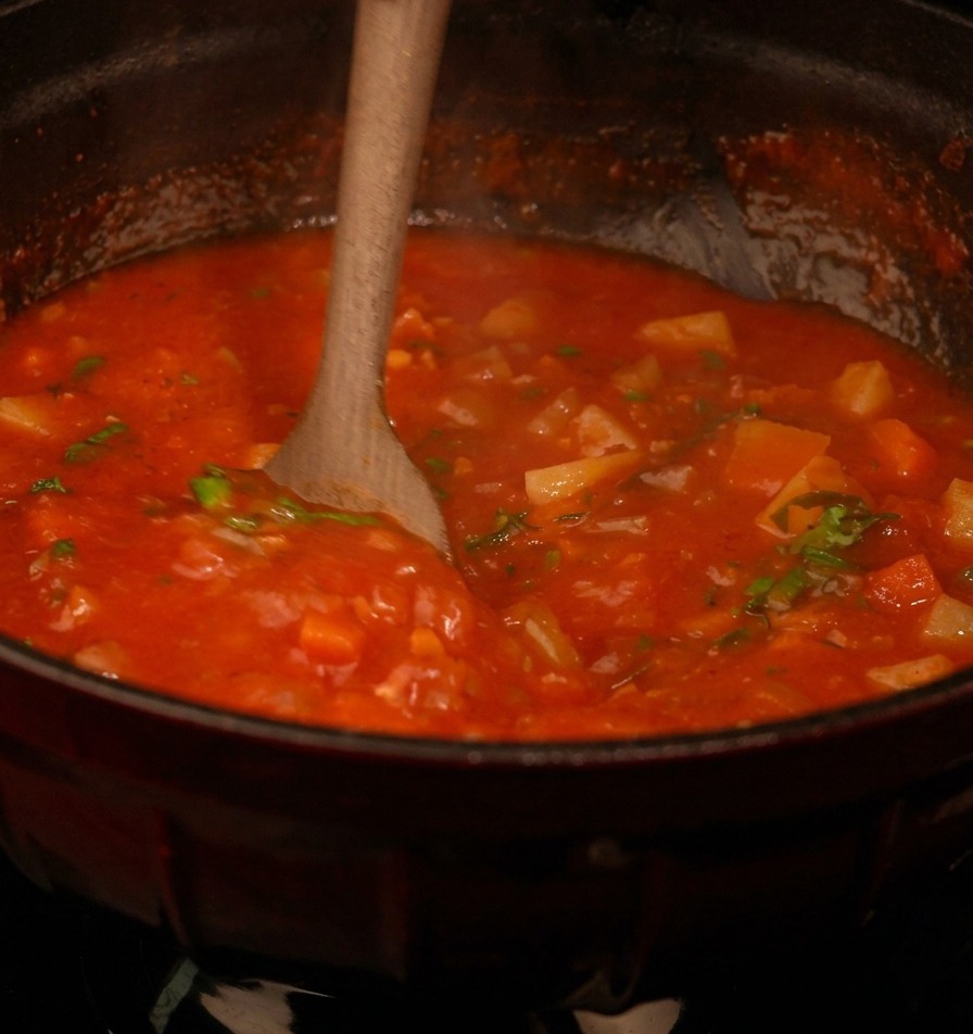 Una cuchara de madera removiendo una sopa espesa a base de tomate rojo llena de verduras en cubitos y motas de perejil verde fresco.
