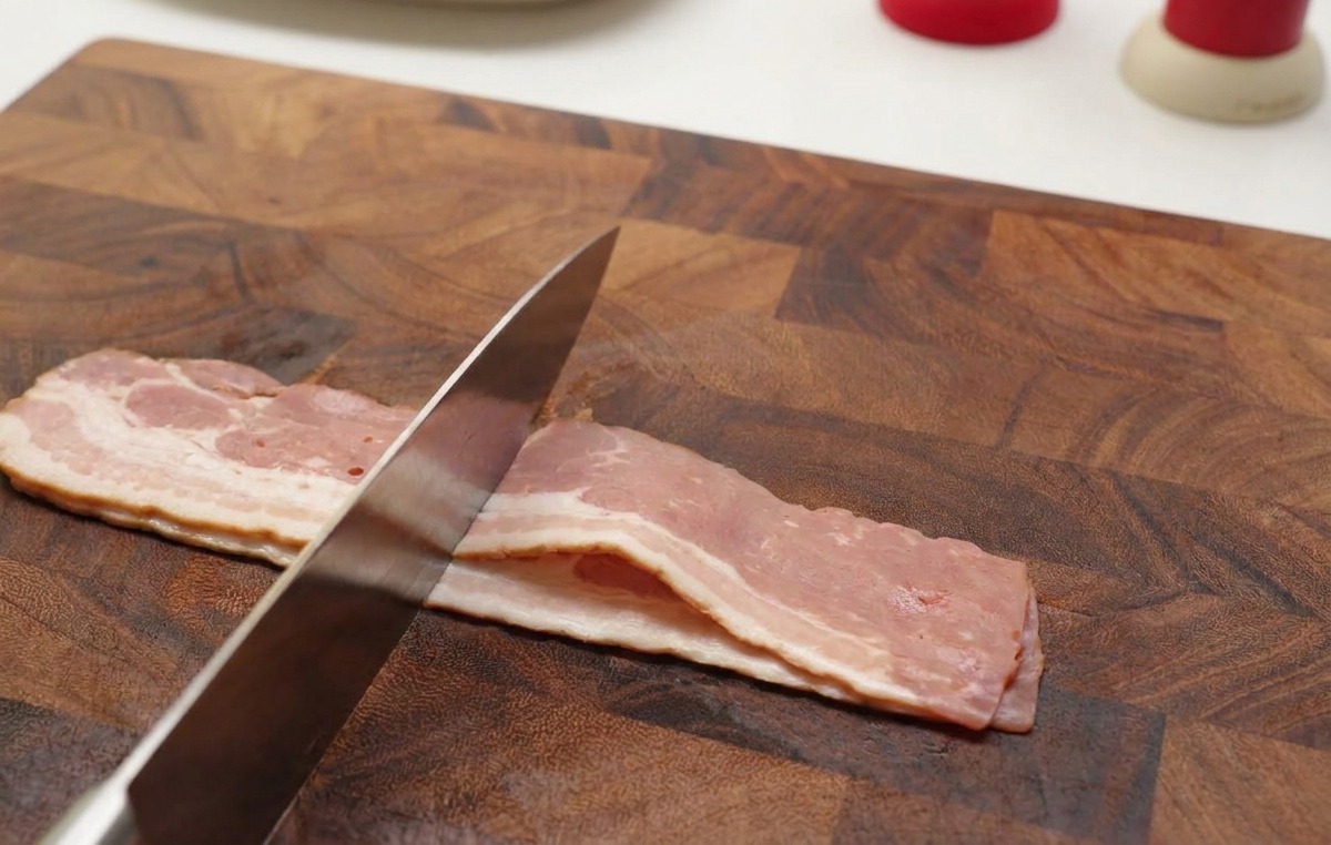 Manos usando un cuchillo afilado para cortar tiras de bacon crudo en trozos pequeños sobre una tabla de madera.
