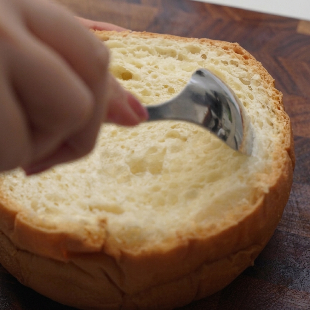 Una mano usando una cuchara metálica para extraer la miga del interior de un pan redondo.