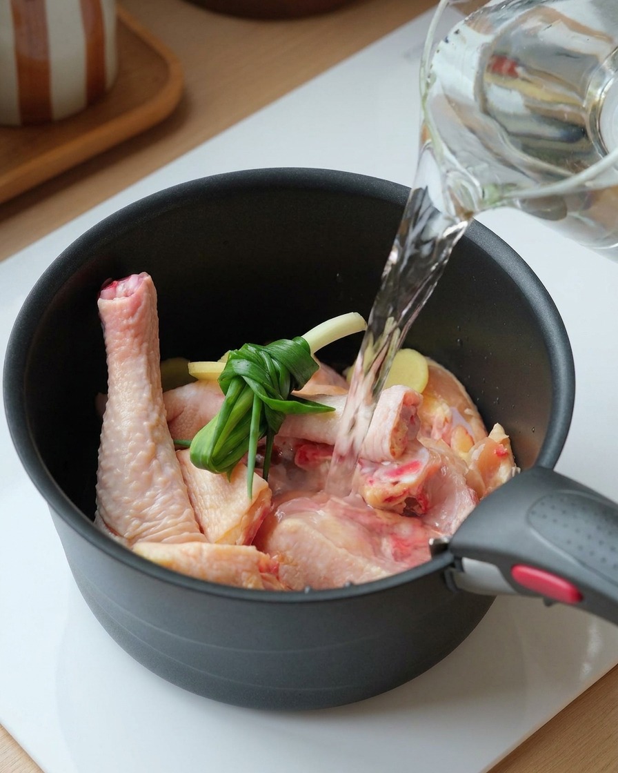 Agua vertiéndose desde una jarra de cristal en una olla con pollo crudo, cebolletas atadas y rodajas de jengibre.