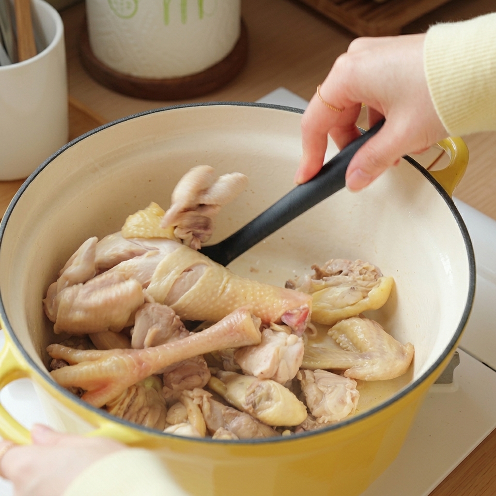 Una mano usando una espátula negra para saltear trozos de pollo blanqueado en una olla amarilla.
