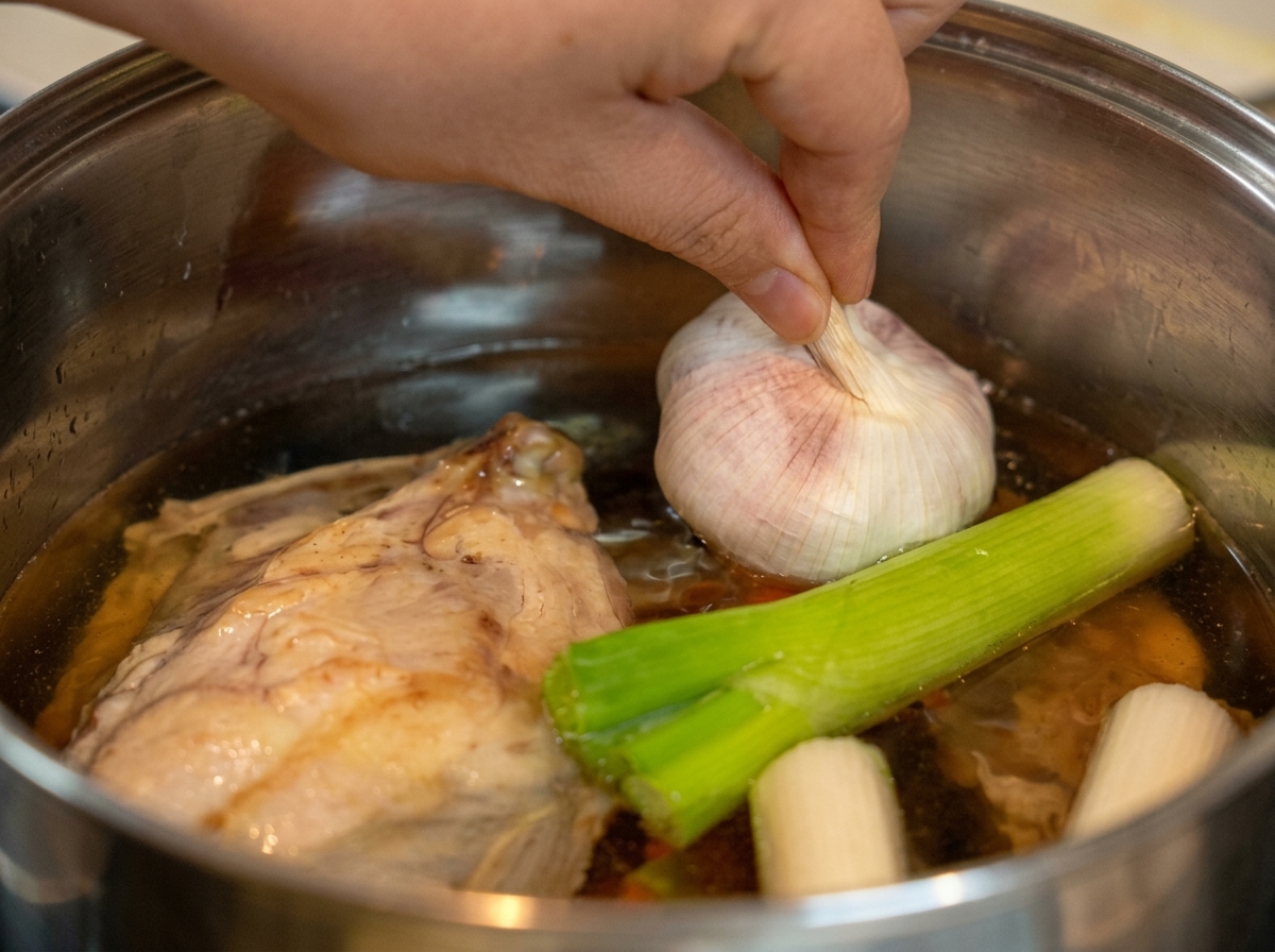Una mano colocando una cabeza de ajo en la olla con el pollo y el puerro.