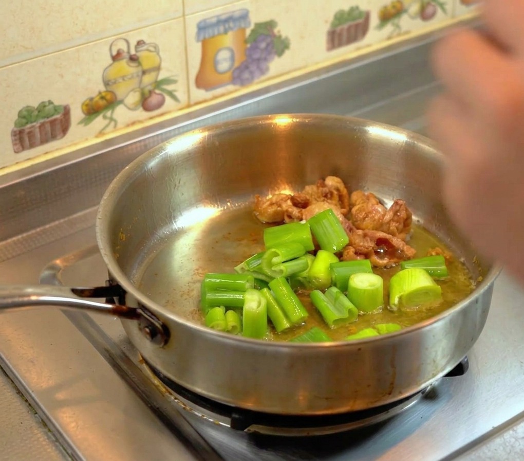 Hojas verdes de puerro friéndose en grasa de pollo dorada en una sartén.