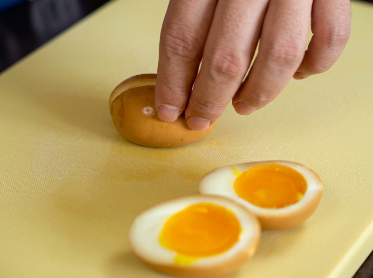 Una mano cortando un huevo marinado por la mitad revelando una yema naranja brillante.