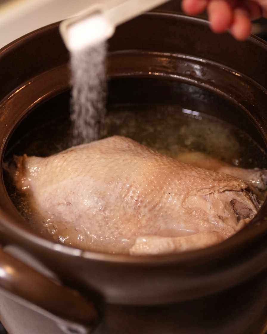 Una mano espolvoreando sal con una cuchara medidora sobre el pollo en el caldo claro.
