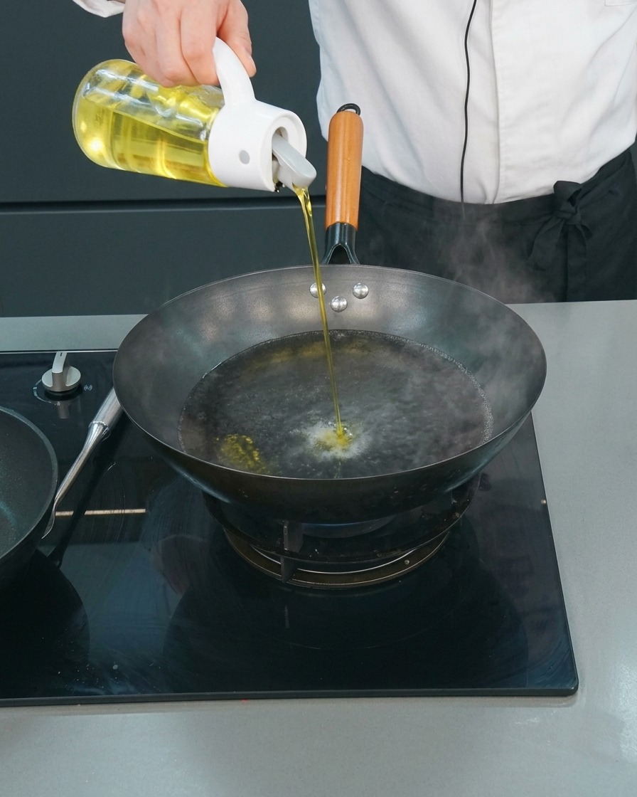 Un chef vertiendo un chorrito de aceite de cocina desde un dispensador de cristal en un wok con agua hirviendo.