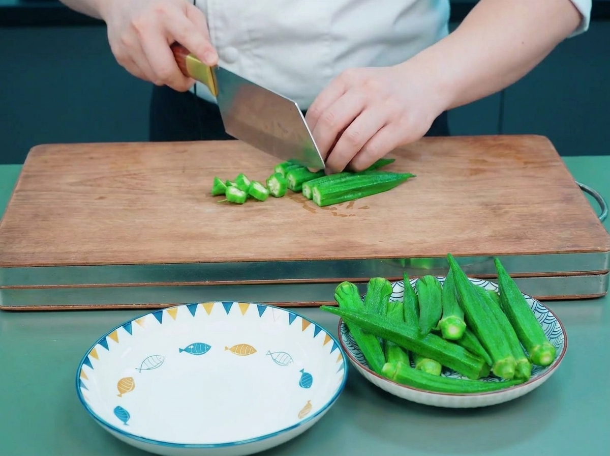 Un chef usando un hacha de cocina para cortar okra escaldada en rodajas con forma de estrella sobre una tabla de madera.