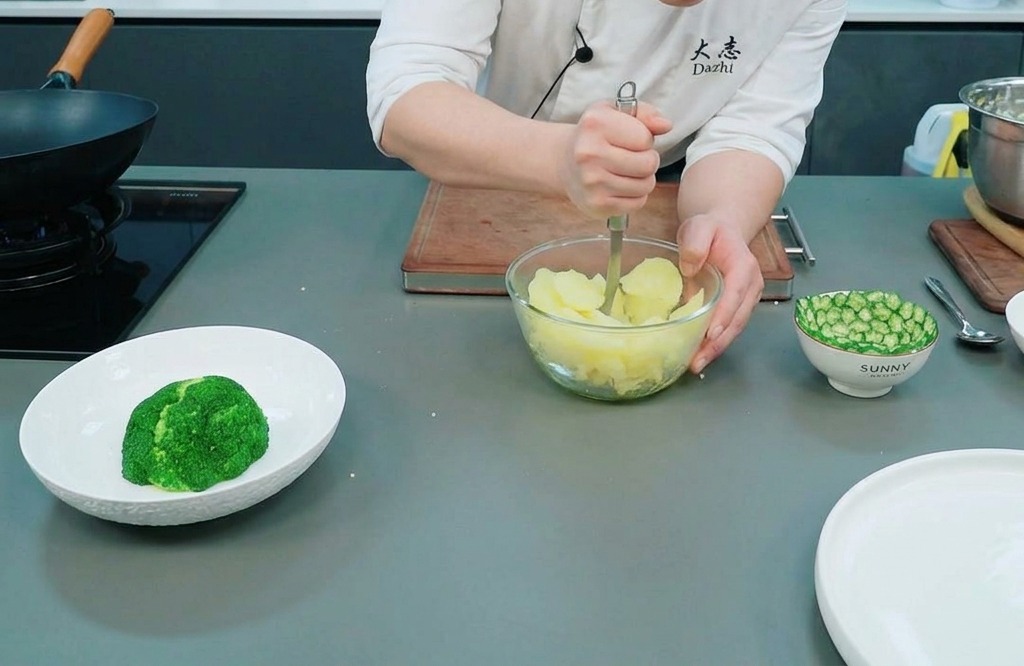 Chef usando un machacador de metal para aplastar patatas al vapor en un bol de cristal sobre el mostrador.