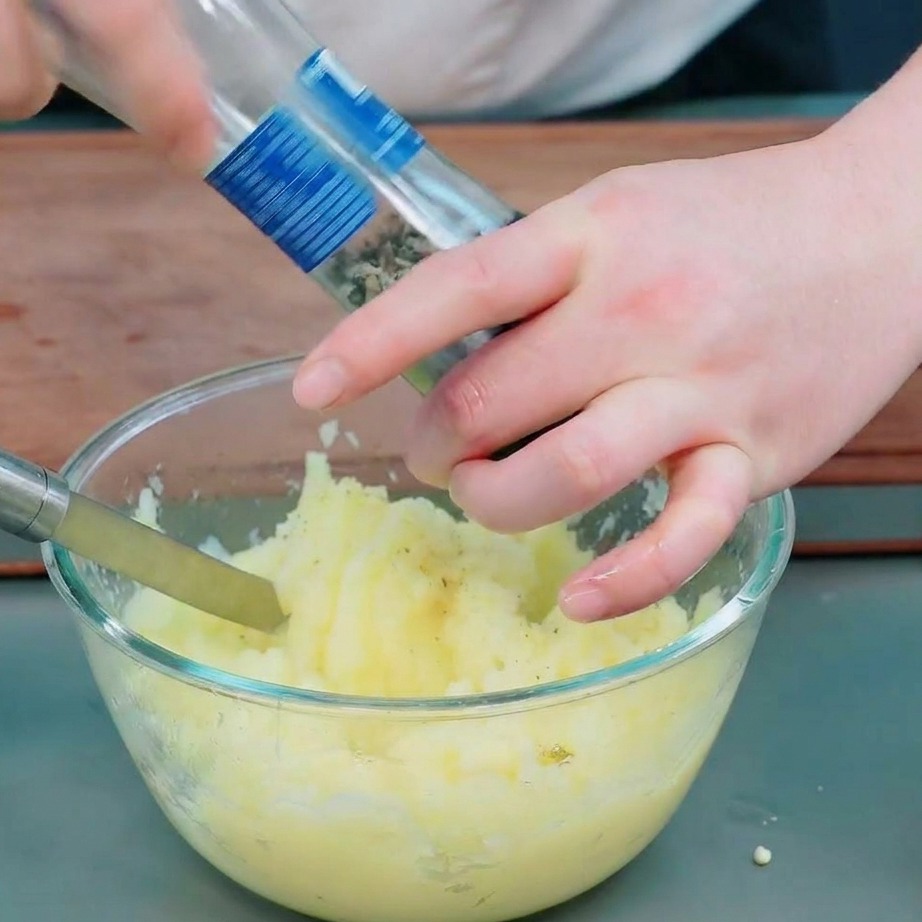 Manos usando un molinillo de cristal para añadir pimienta negra y sal al puré de patatas suave.