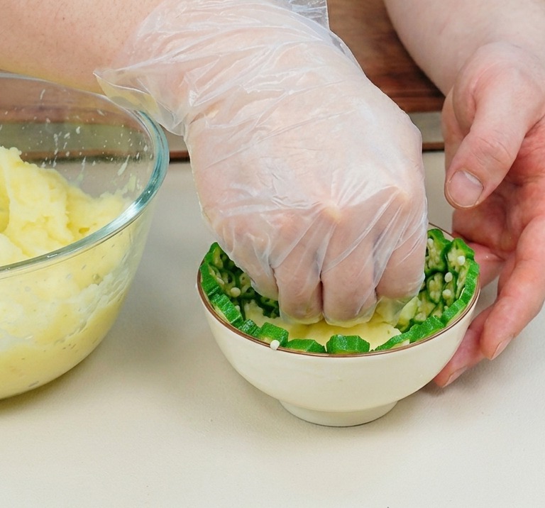Una mano con guante de plástico presionando puré de patatas en un bol pequeño forrado con rodajas de okra.