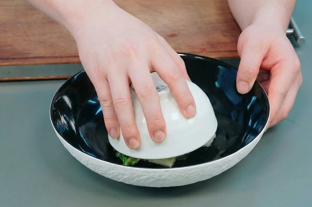 Manos levantando con cuidado un bol blanco pequeño de un bol negro grande para revelar el domo de okra y patata.