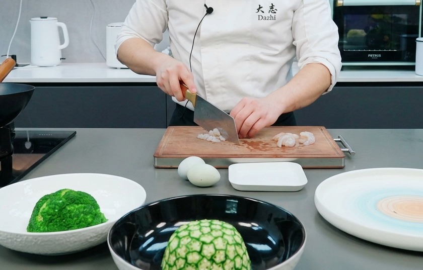 Un chef usando un hacha de cocina para picar gambas crudas en trozos pequeños sobre una tabla de madera.