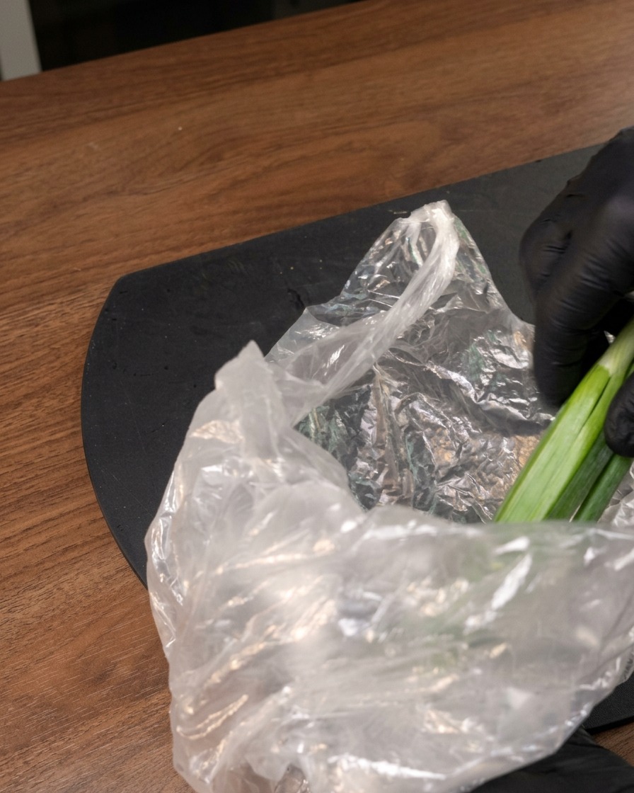 Una mano con guante sacando vegetales verdes frescos de una bolsa de plástico transparente.