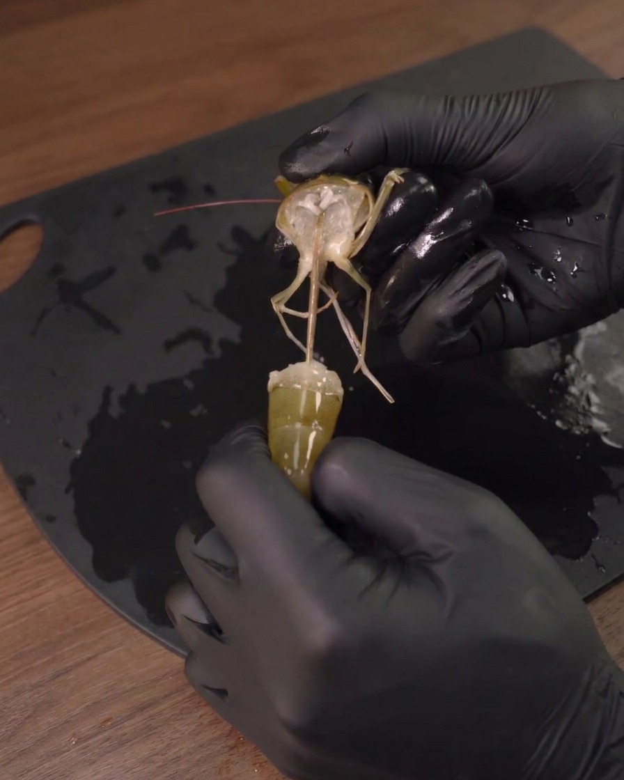 Una persona retirando la cabeza de una gamba cruda con guantes negros.