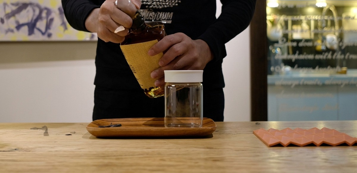 Manos vertiendo bourbon desde una botella a un frasco de vidrio transparente sobre una mesa de madera.