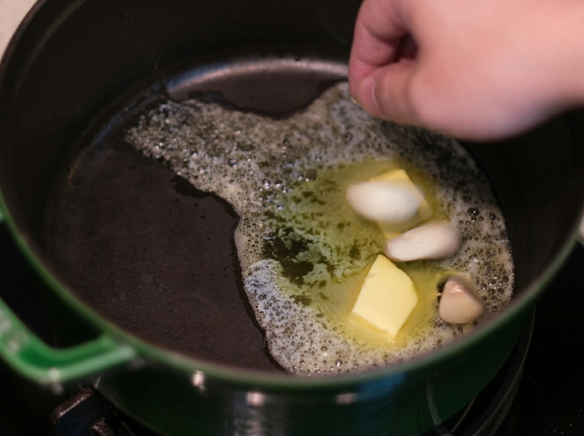 Una mano añadiendo dientes de ajo a la mantequilla derretida dentro de una olla de hierro verde.