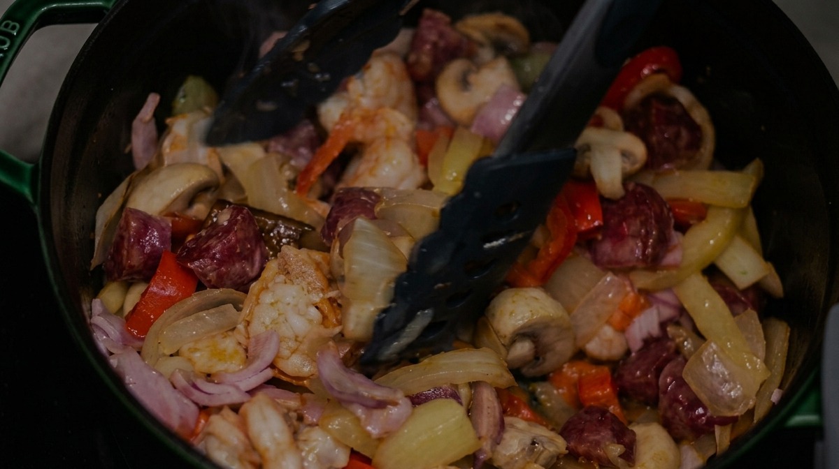 Mezclando salchicha ahumada, tocino y camarones con los vegetales usando pinzas negras.
