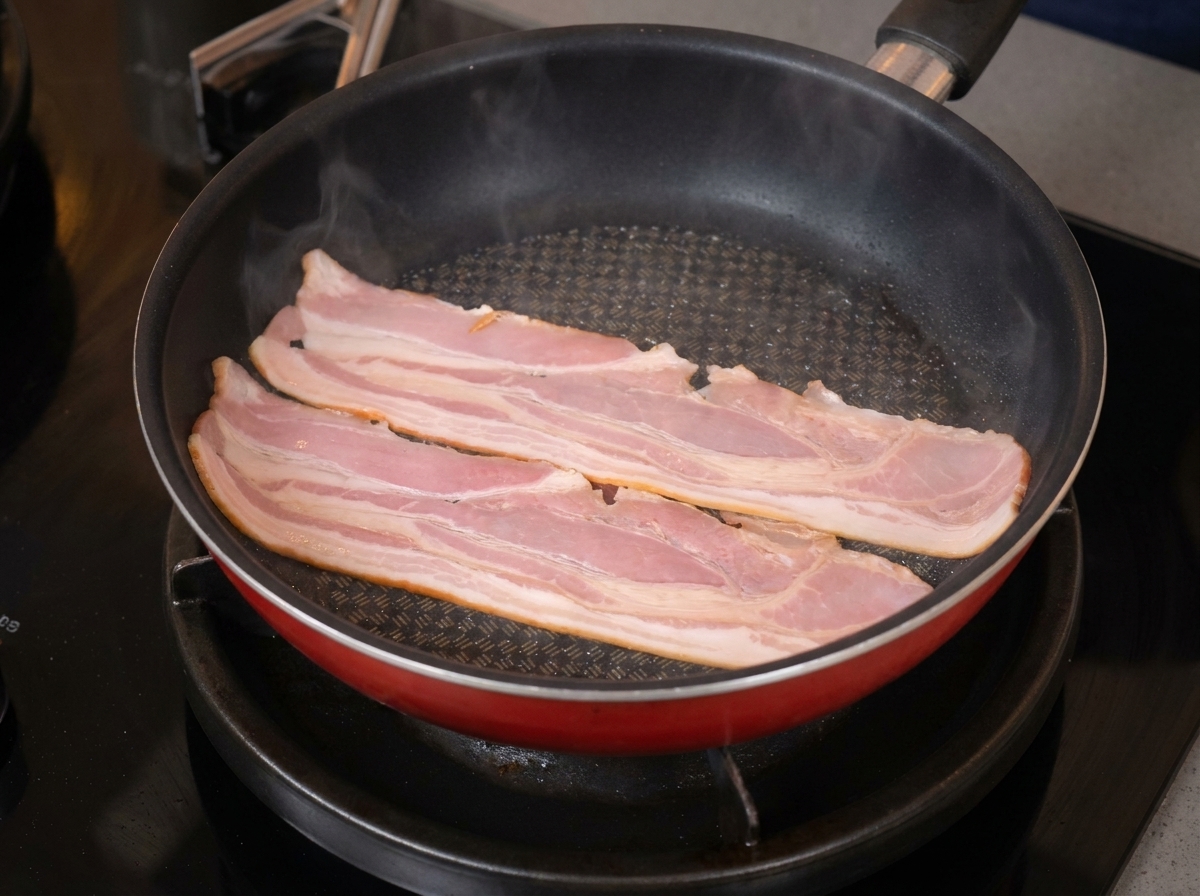 Dos tiras largas de tocino burbujeando y empezando a encogerse en una sartén antiadherente roja.