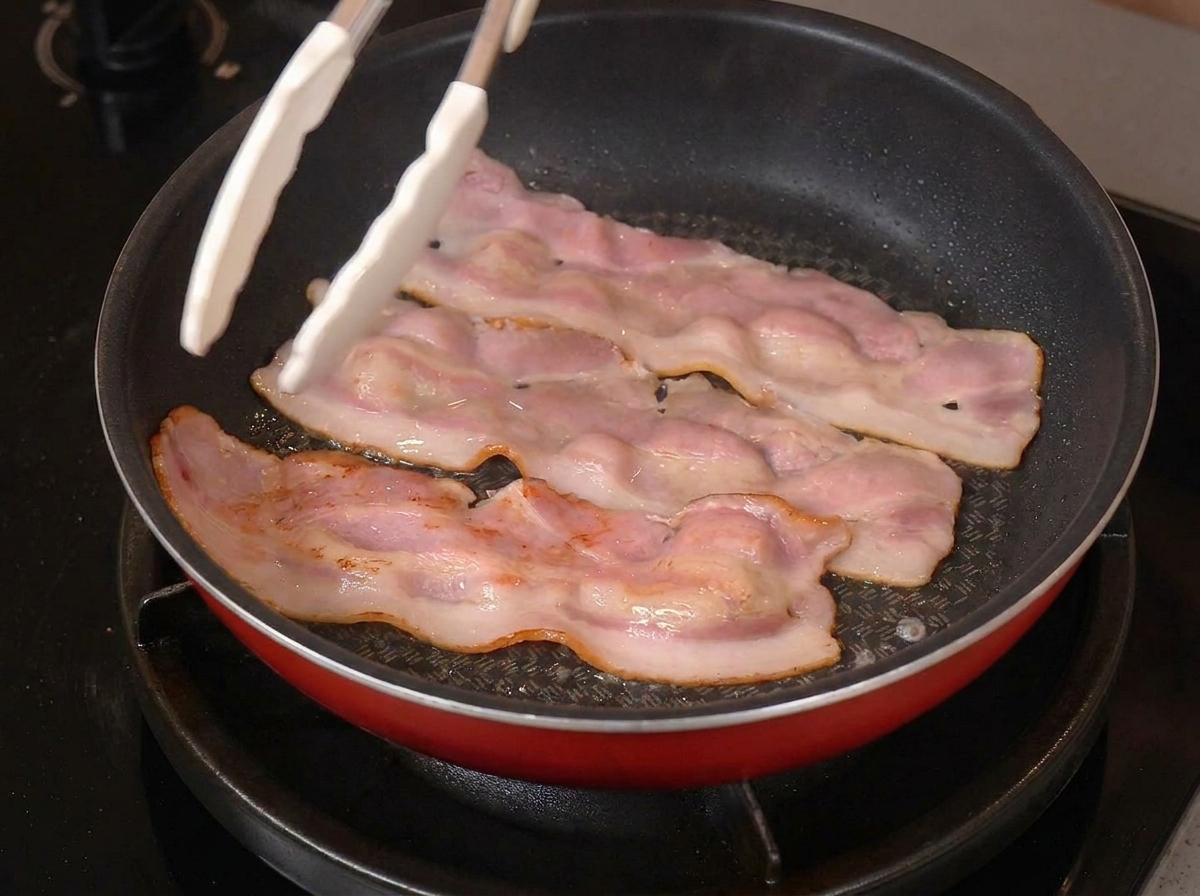 Tres tiras de tocino chisporroteando en una sartén con los bordes dorados y una pinza blanca sujetando una.
