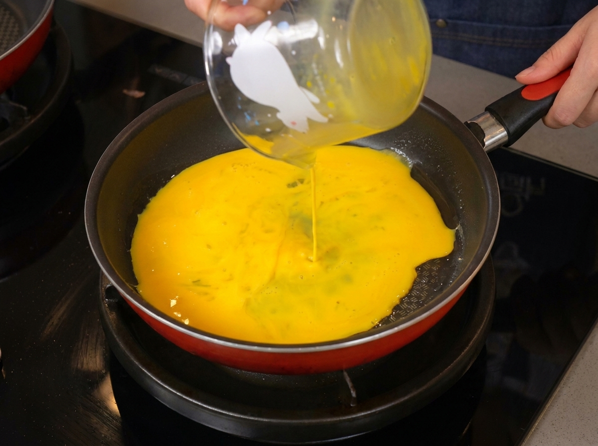 Huevos batidos de color amarillo brillante vertiéndose desde una jarra de vidrio a una sartén caliente.