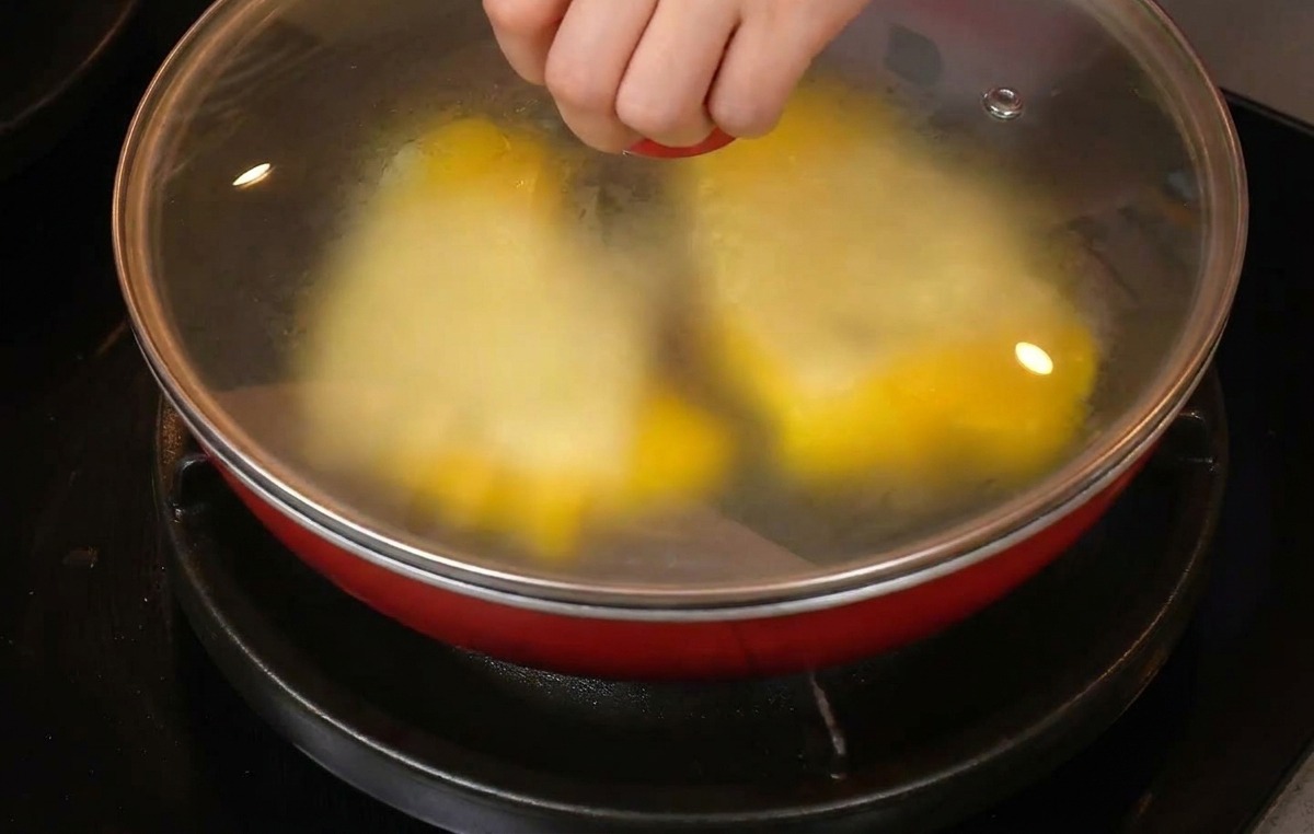 Una mano colocando una tapa de vidrio en una sartén roja con huevos revueltos y rebanadas de queso blanco.