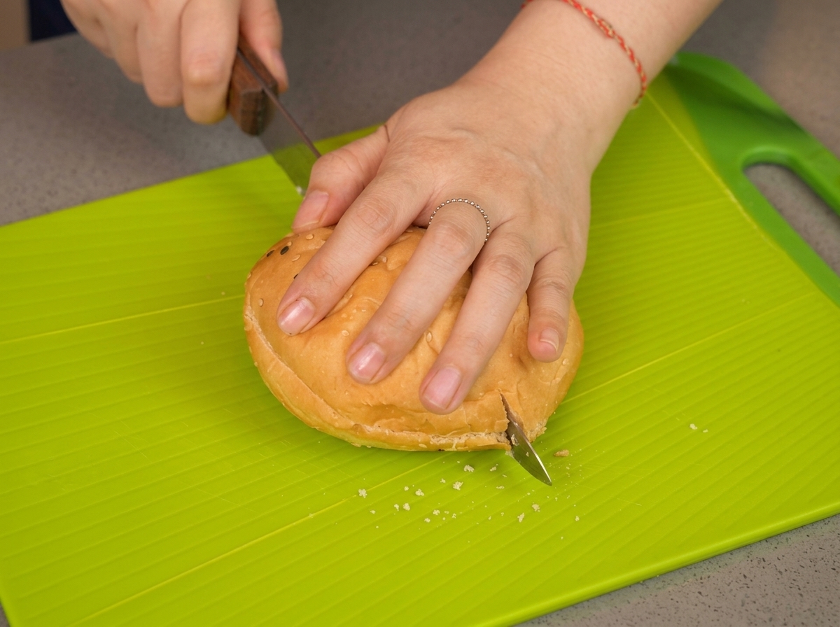 La mano de una persona presionando un pan de sésamo mientras un cuchillo lo corta por la mitad en una tabla verde.