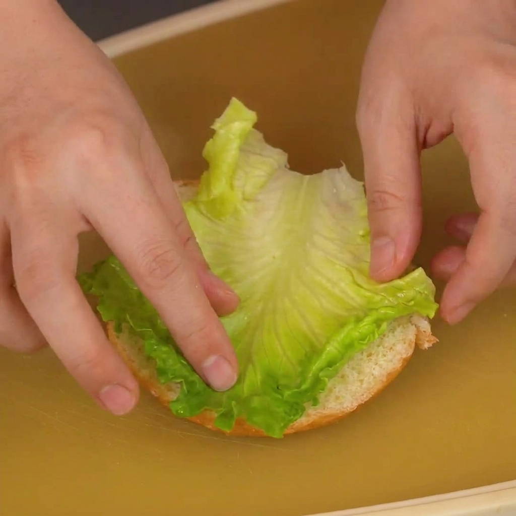 Manos colocando una hoja de lechuga verde vibrante sobre la base de un pan de hamburguesa.