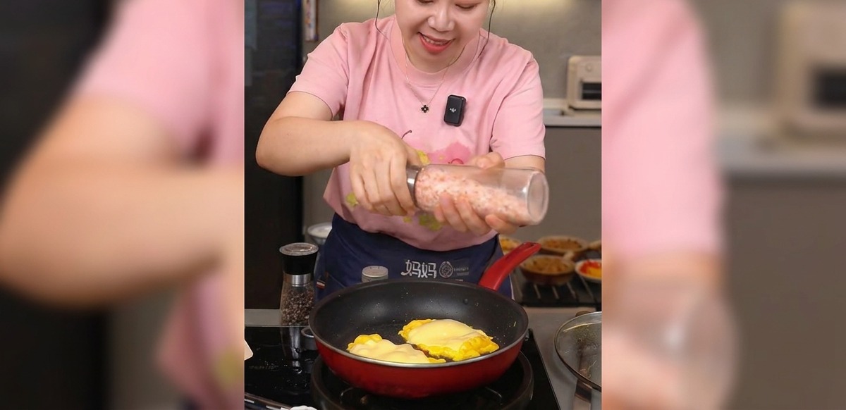 Manos usando un molinillo para esparcir sal marina rosa sobre una sartén con huevos cocinados y queso derretido.