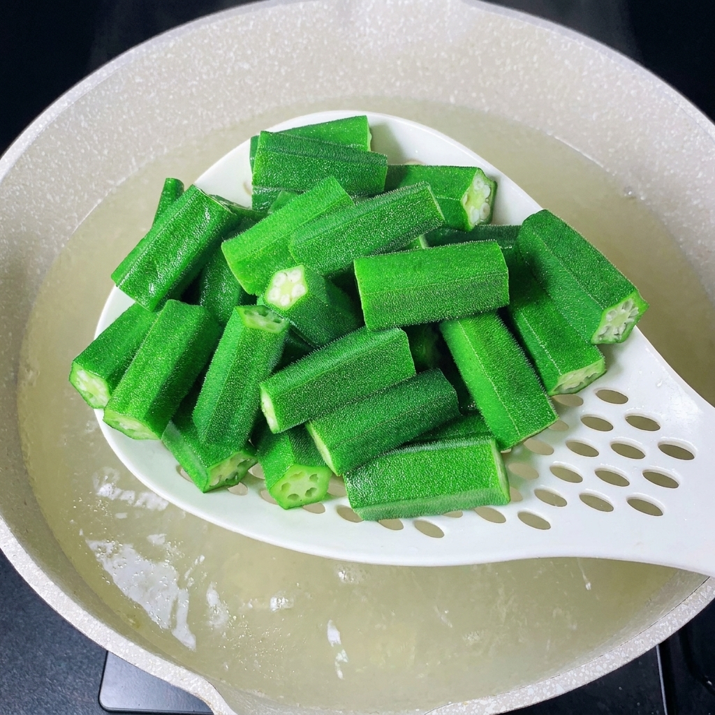Una espumadera retirando la okra mientras se añaden jengibre y cebolleta al agua.