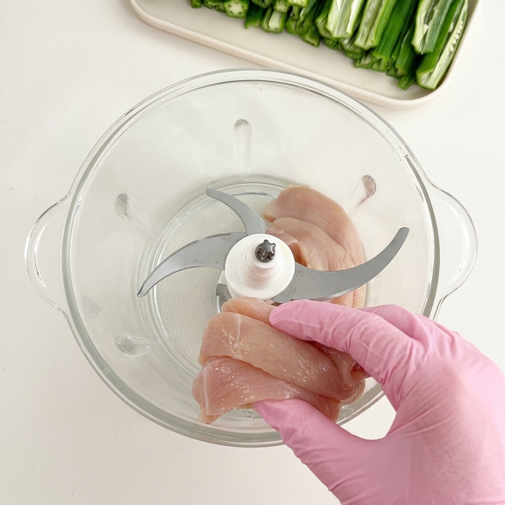 Mano con guante introduciendo trozos de pechuga de pollo cruda en el bol de cristal de un procesador de alimentos.