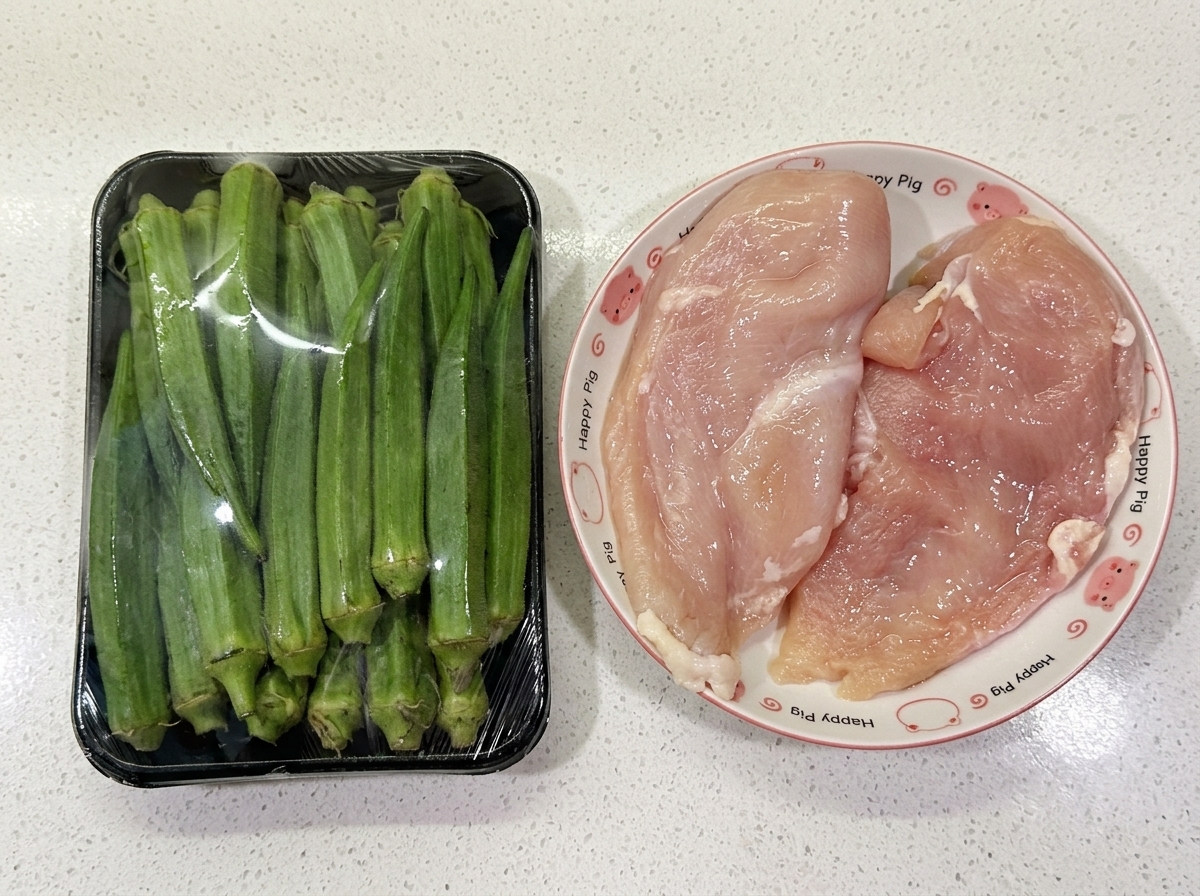 Un paquete de okra verde fresca junto a dos pechugas de pollo crudas en un plato sobre un mostrador blanco.