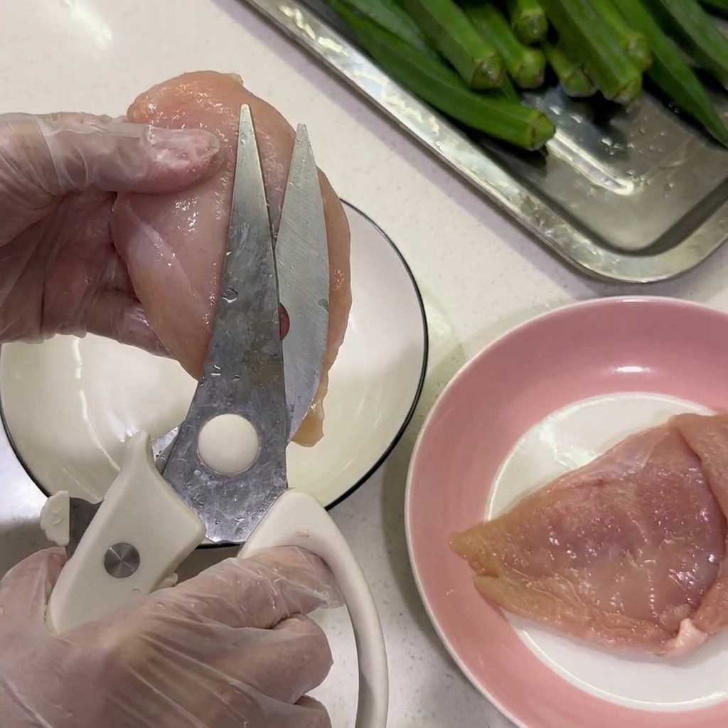 Manos con guantes usando tijeras de cocina para recortar la grasa blanca de una pechuga de pollo cruda.