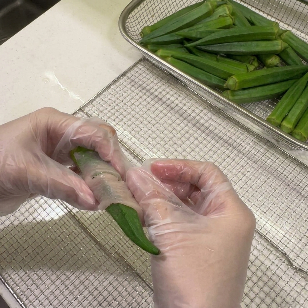 Manos envolviendo firmemente una tira fina de pollo crudo alrededor de una vaina de okra sobre una rejilla de deshidratador.
