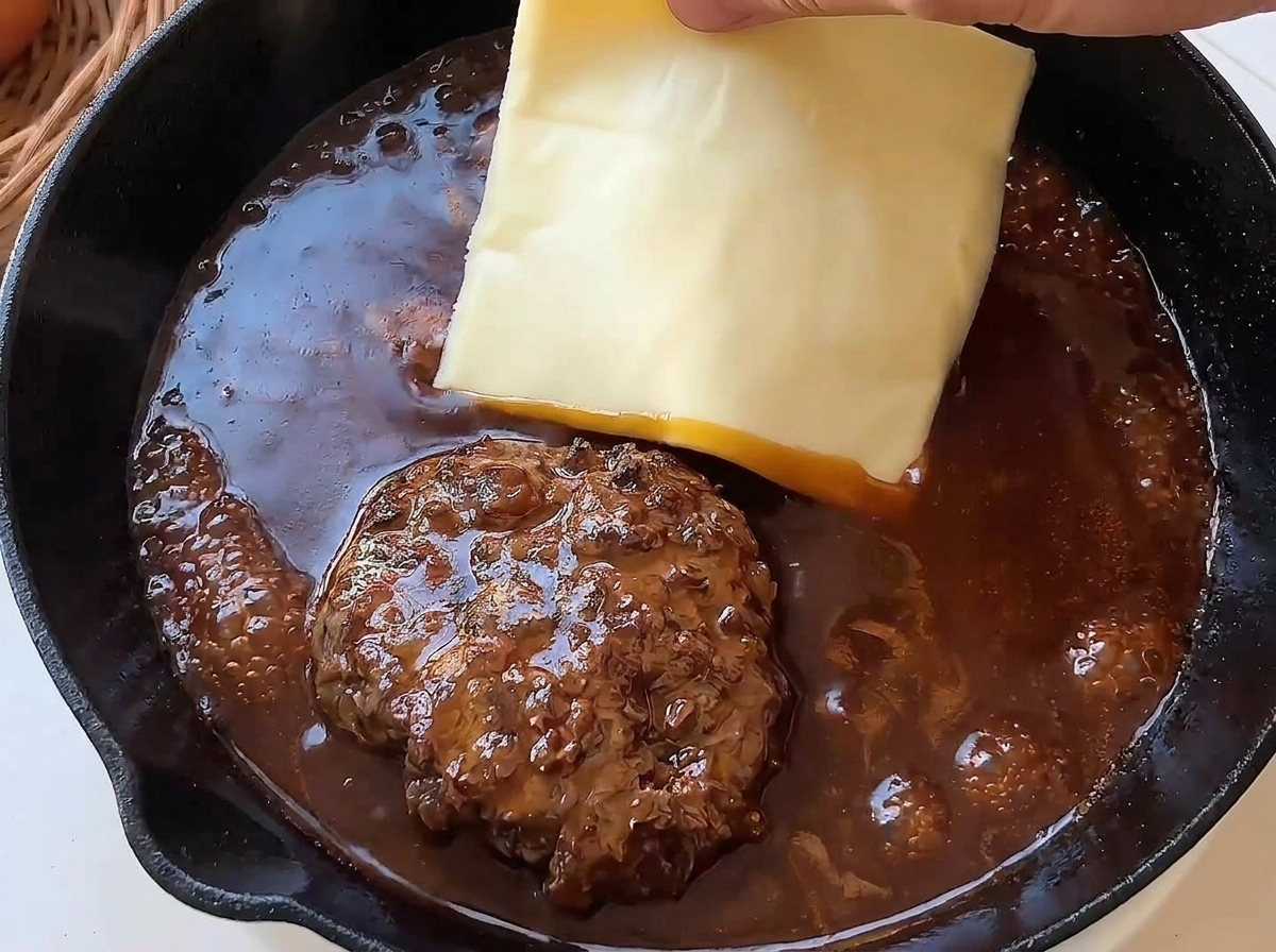 Una mano colocando una loncha de queso amarillo sobre una hamburguesa en la sartén.