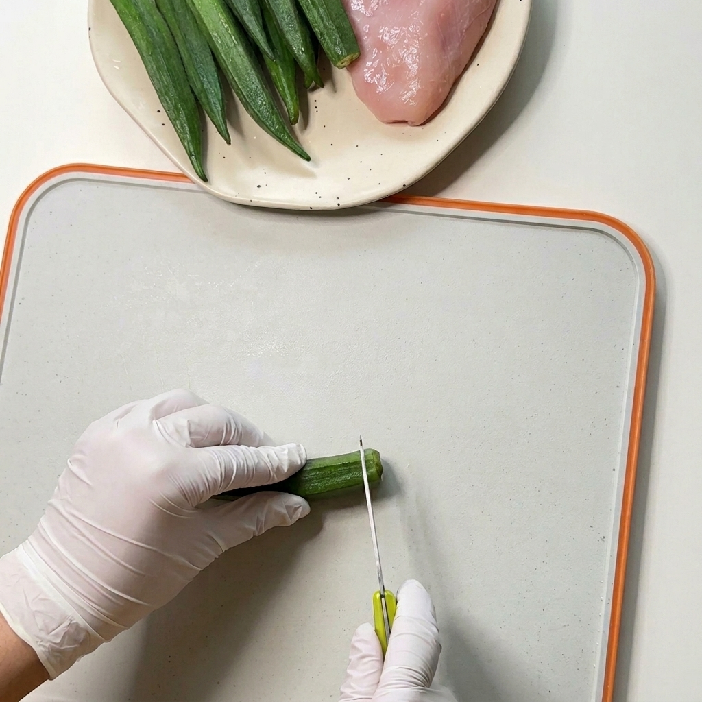 Manos con guantes usando un cuchillo pequeño para cortar los extremos de okras verdes frescas en una tabla de cortar blanca.