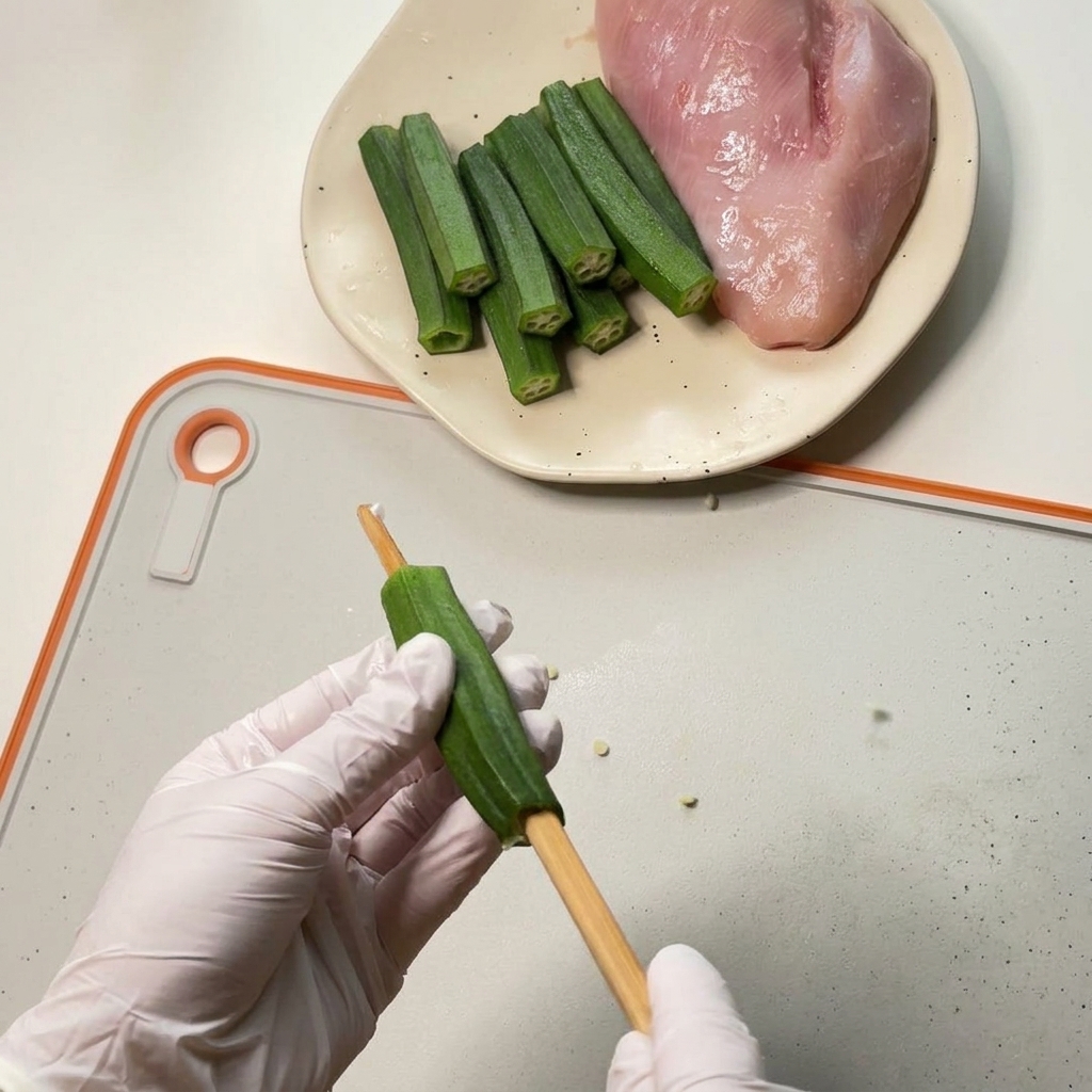 Manos con guantes ahuecando el centro de una okra verde usando un palillo de madera grueso sobre una tabla de cortar blanca.