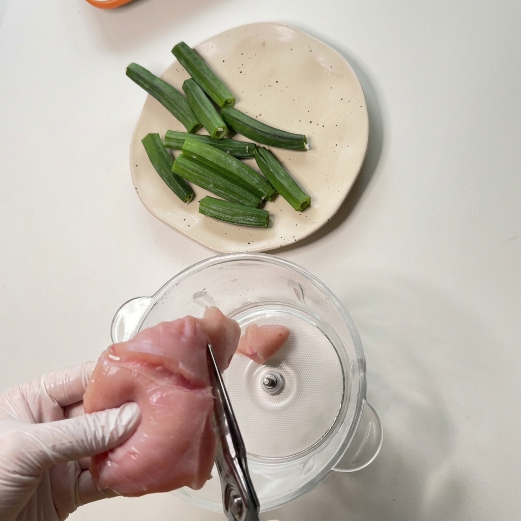 Manos con guantes usando tijeras de cocina para cortar un trozo de pechuga de pollo cruda sobre el vaso de cristal de un procesador de alimentos.