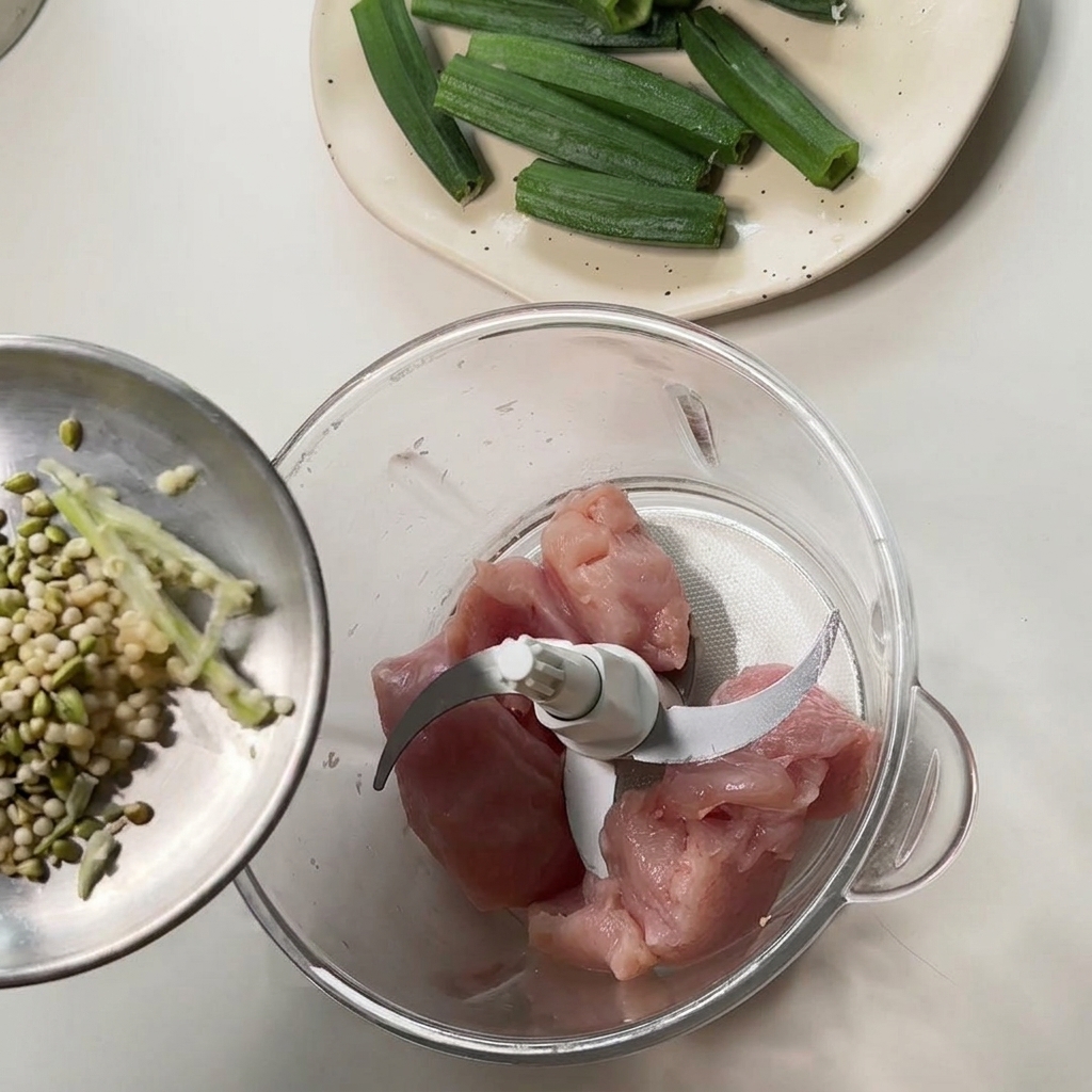 Vertiendo semillas y restos de okra desde un pequeño recipiente de metal en un procesador de alimentos que contiene trozos de pollo crudo.