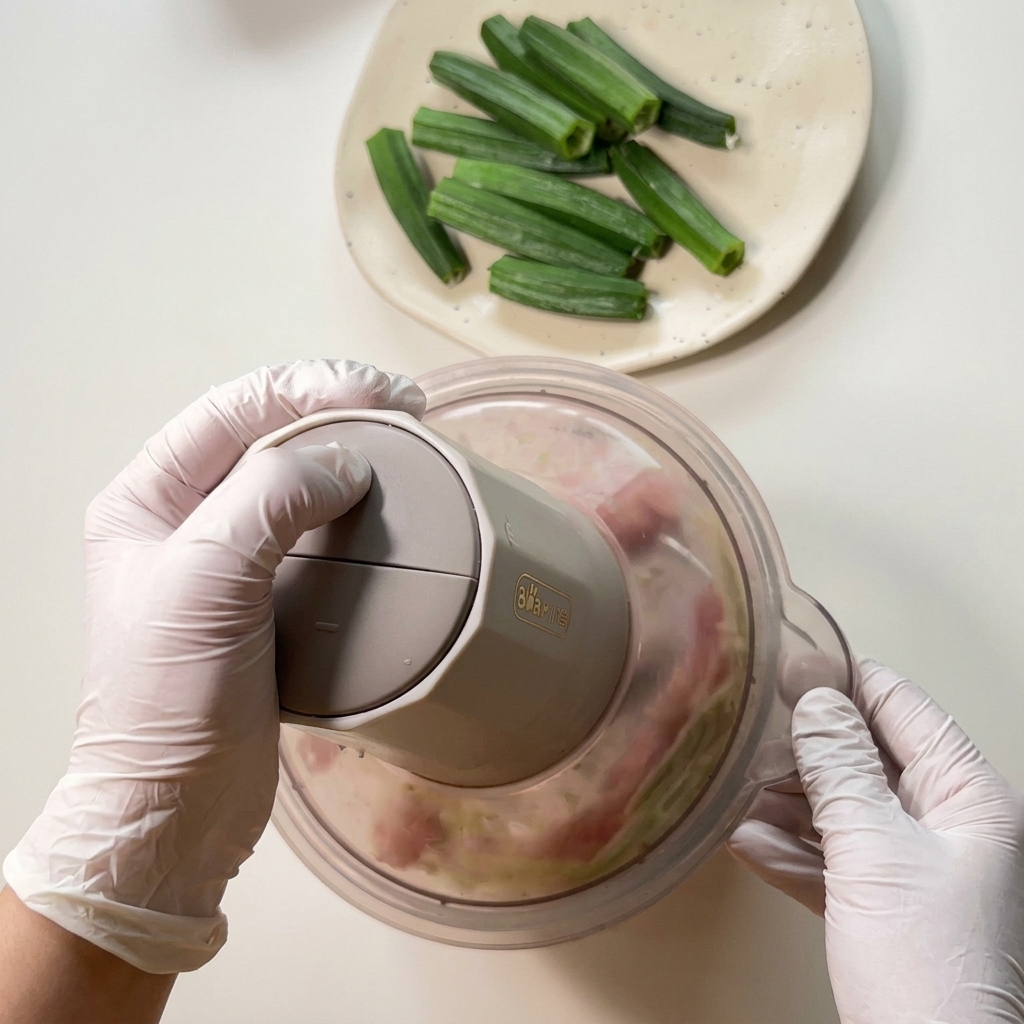 Manos con guantes presionando un mini procesador de alimentos para triturar pollo crudo y restos de okra hasta formar una pasta.