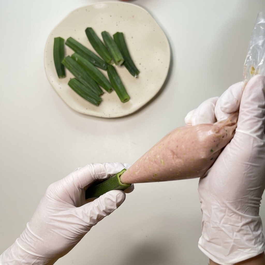 Manos con guantes usando una manga pastelera de plástico para inyectar un relleno de pollo picado en una vaina de okra ahuecada.