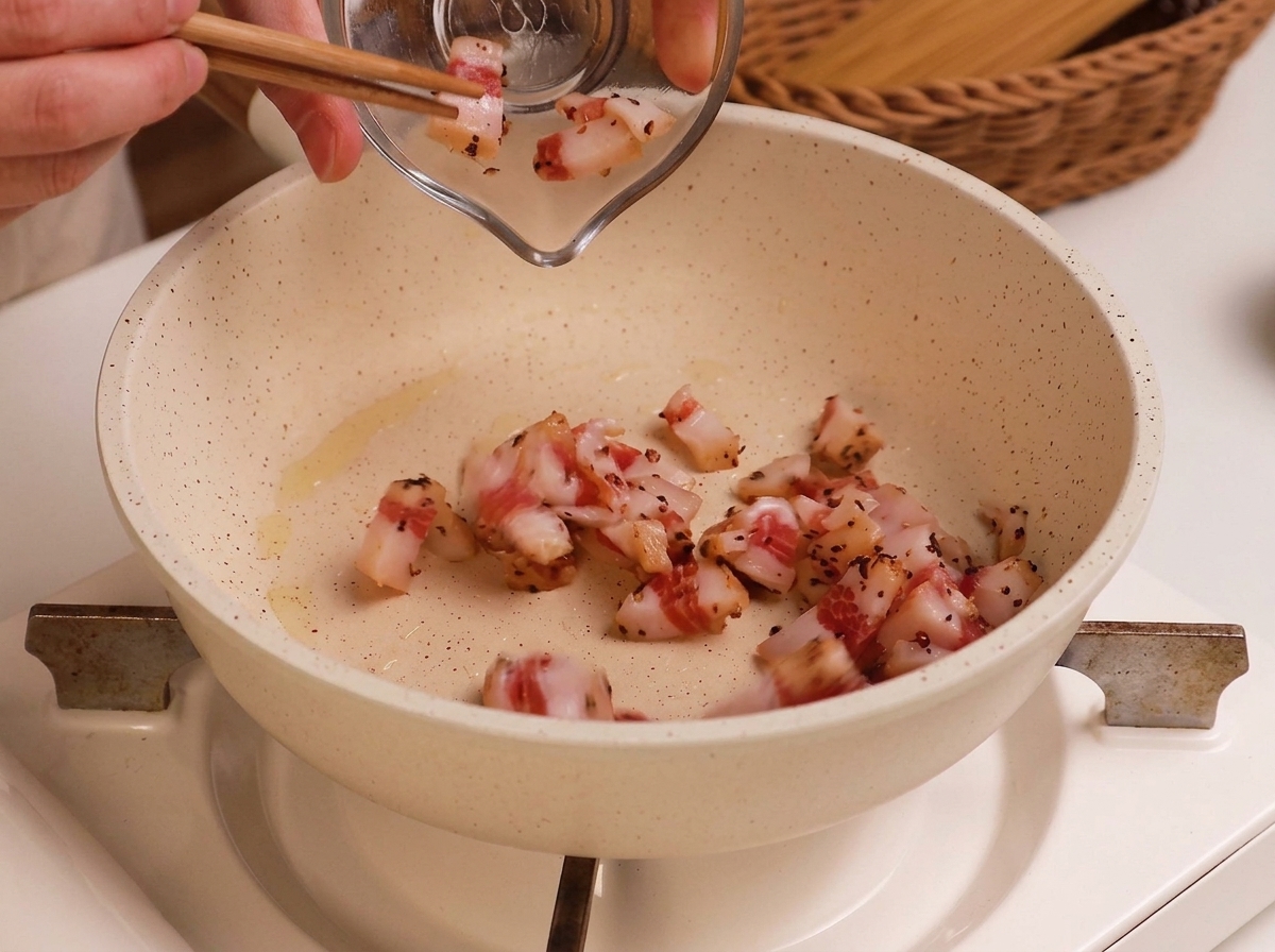 Guanciale troceado pasando de un bol de cristal a una sartén blanca ligeramente aceitada.