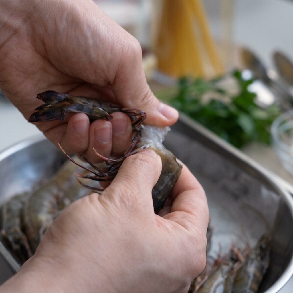 Manos pelando un langostino crudo sobre un bol de acero inoxidable, conservando la cabeza.