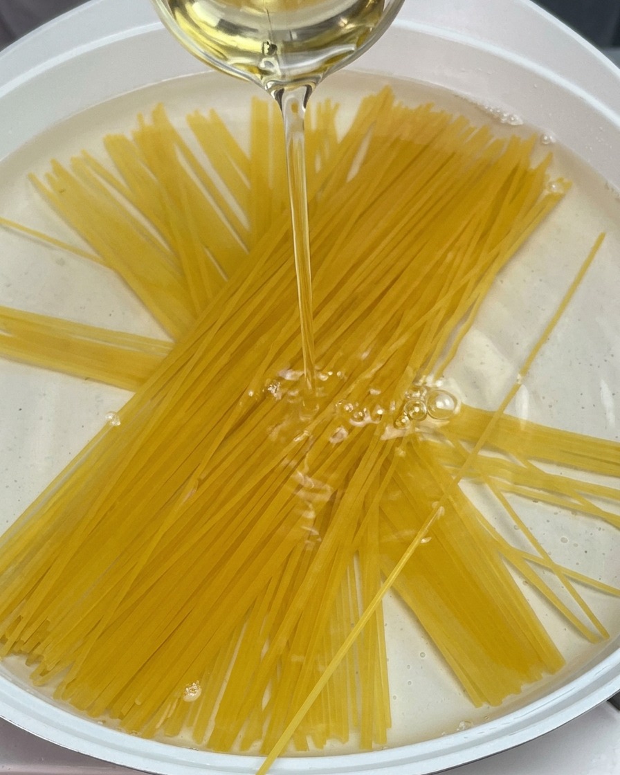 Espaguetis secos sumergidos en una olla con agua mientras cae un chorrito de aceite.