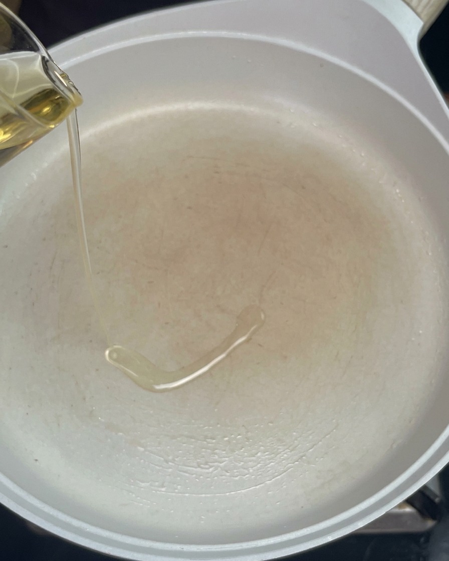 Aceite de cocina vertiéndose desde una jarrita de cristal a una sartén blanca precalentada.