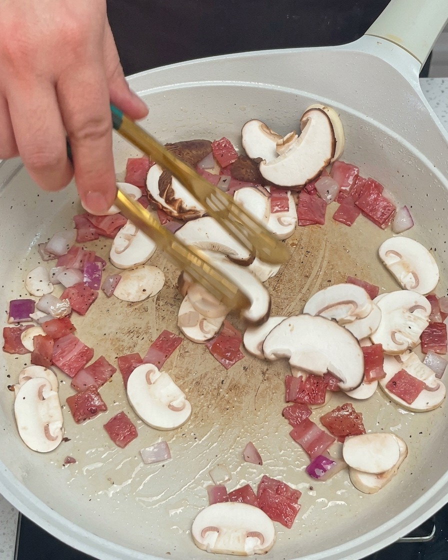 Mano usando pinzas para mezclar setas, cebolla y bacon en una sartén blanca.