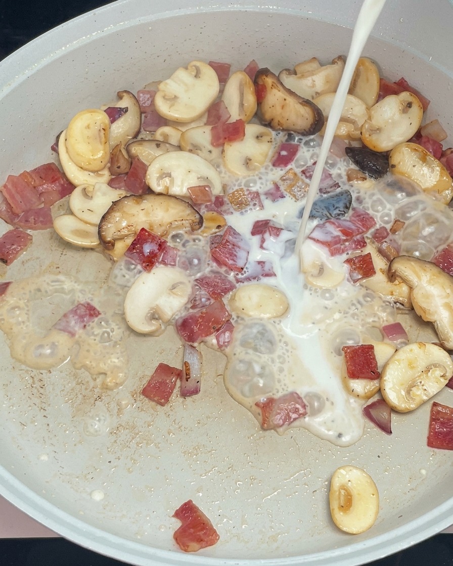 Leche blanca vertiéndose sobre el salteado de champiñones y bacon en la sartén.