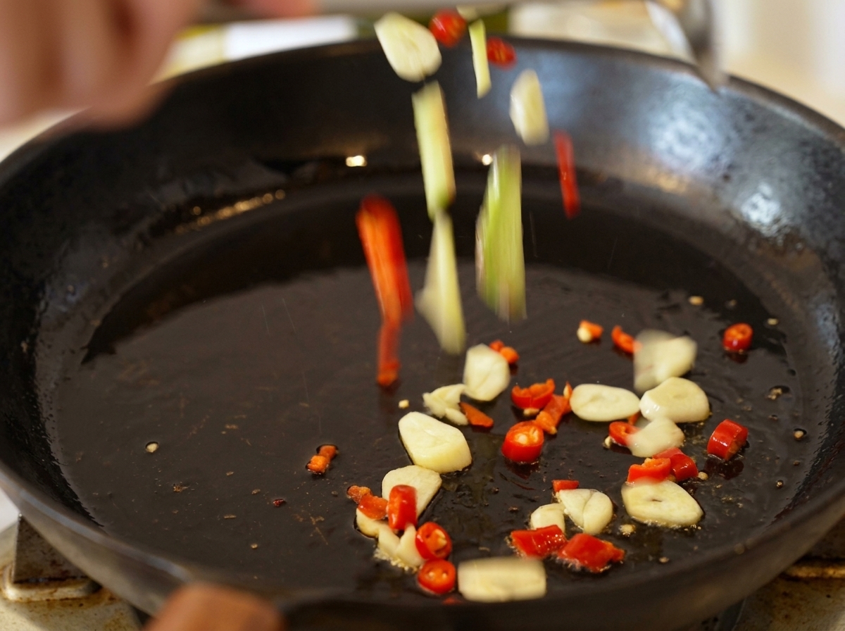Láminas de ajo y trozos de guindilla cayendo en una sartén con aceite de oliva caliente.