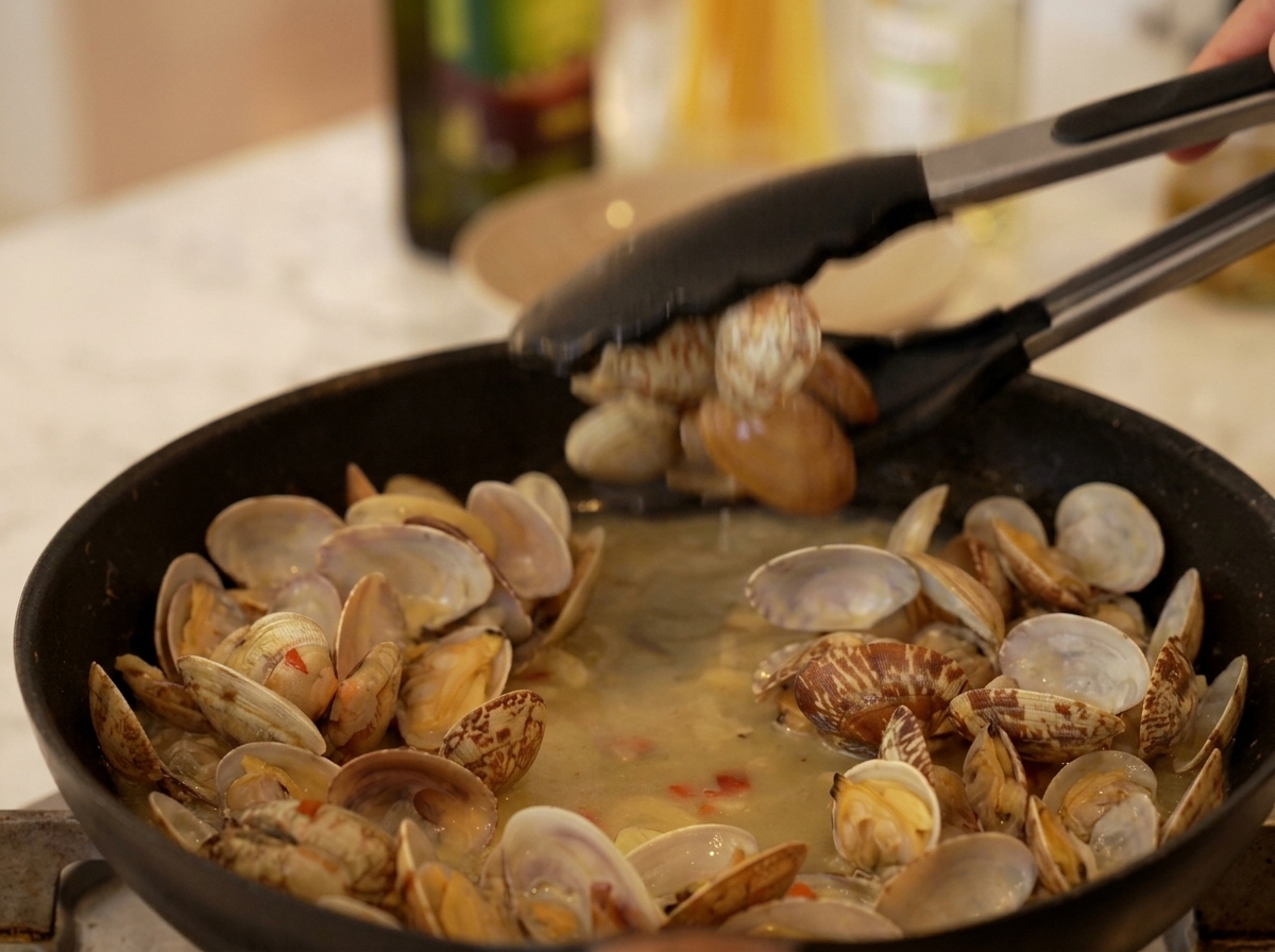 Pinzas de cocina recogiendo almejas abiertas y cocinadas de un caldo en una sartén negra.