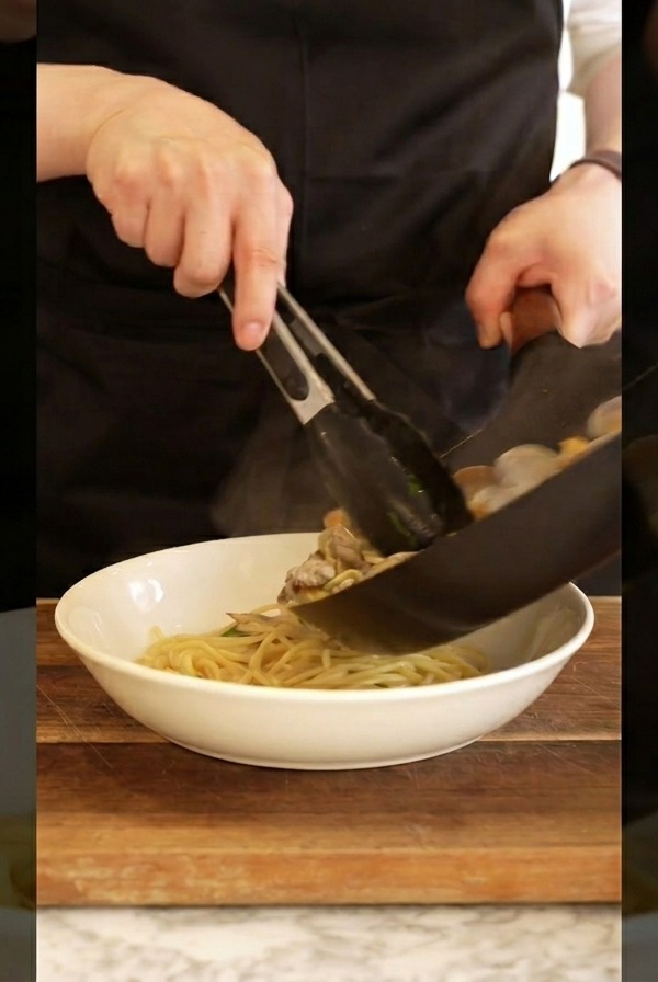 Un cocinero con delantal negro usando pinzas para pasar los espaguetis con almejas de la sartén a un bol blanco.