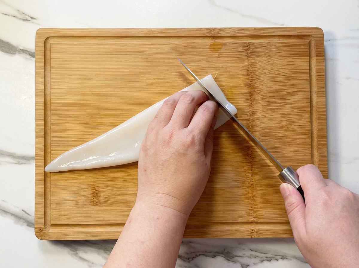 Una persona cortando un tubo de calamar blanco crudo en anillos sobre una tabla de bambú.